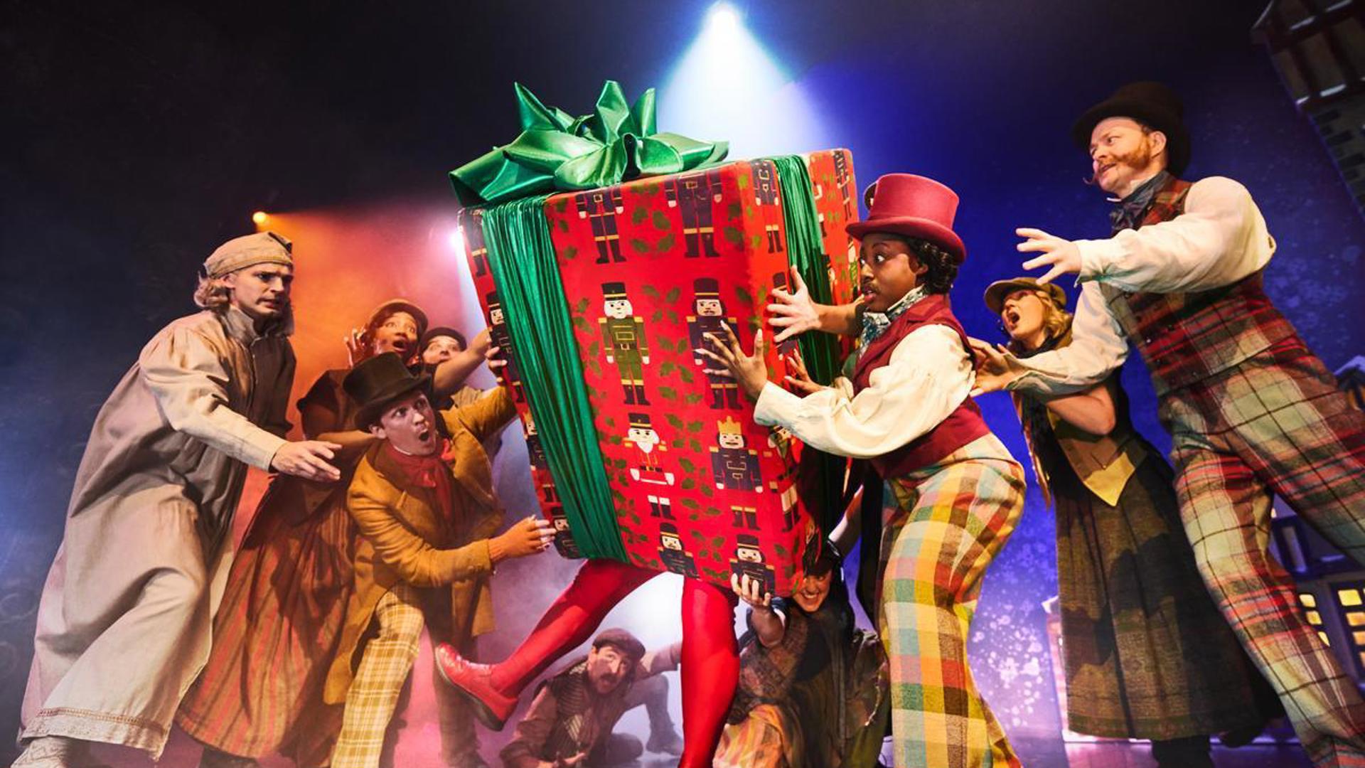 Nine performers are gathered around a large present, holding it up.