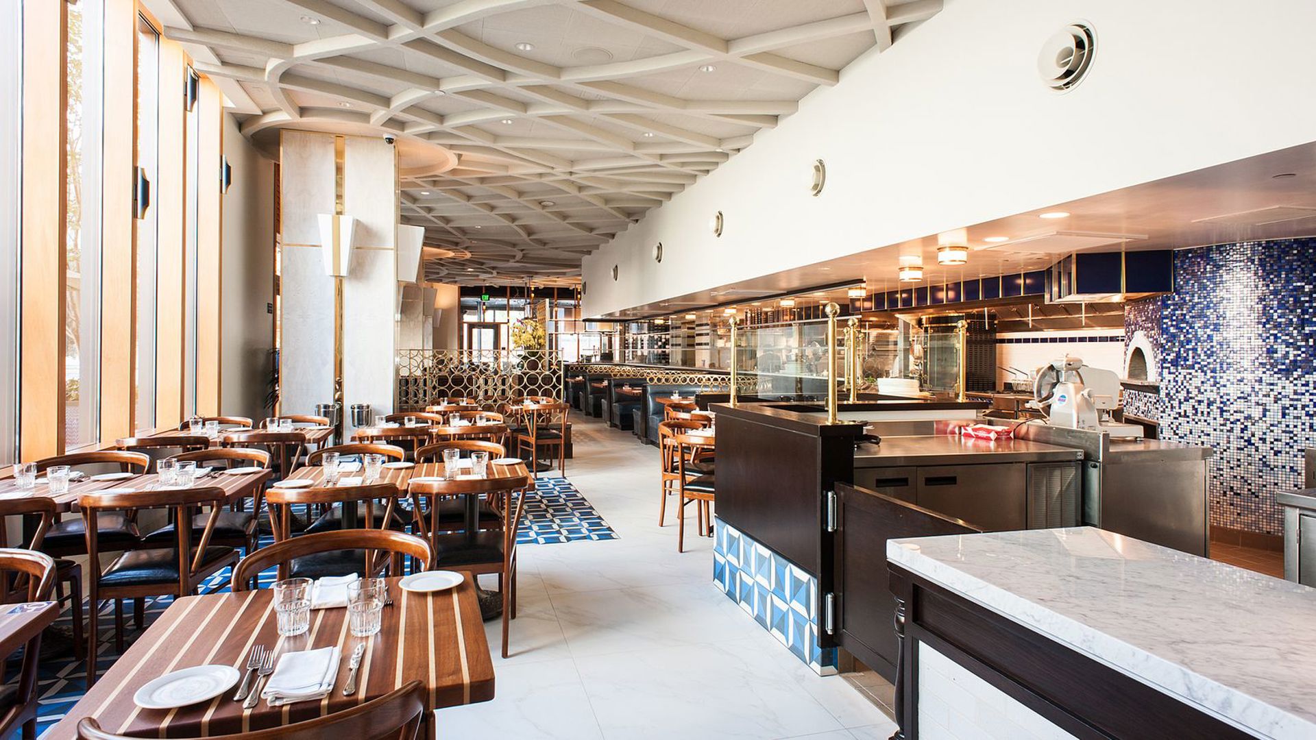 Bright, modern Italian restaurant interior with wood-striped dining tables and chairs on patterned tile floors, a sculptural woven ceiling, floor-to-ceiling windows, and an open kitchen with a blue mosaic-tiled pizza oven in the back.