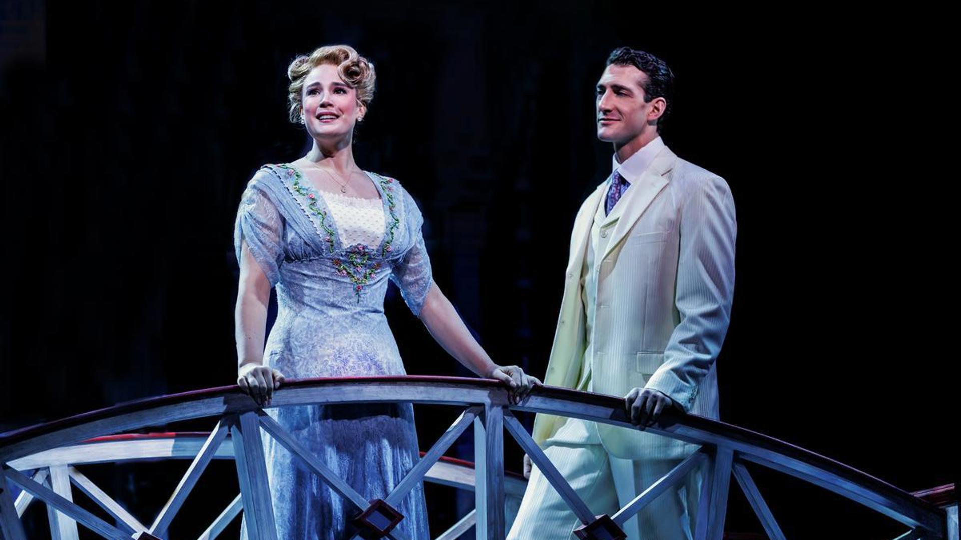 Two actors stand together on a bridge, one looks out at the audience smiling. The other actor looks at the actor smiling.
