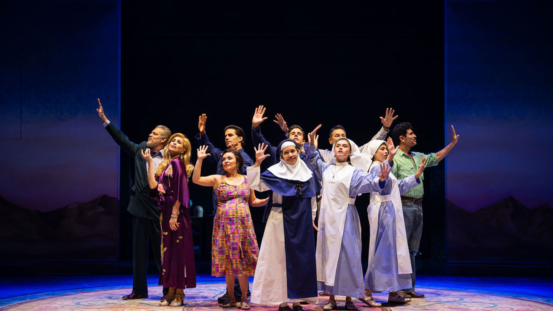 A group of actors stand center stage with their hands up.