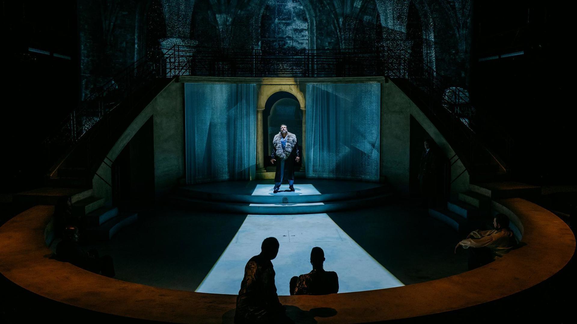 An actor stands center stage with a white light shining over them. Two shadowed actors sit downstage.