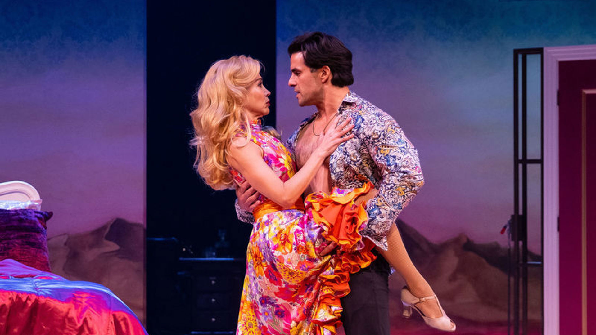 A man and woman stand onstage, embracing. The man holds the woman's leg up romantically.