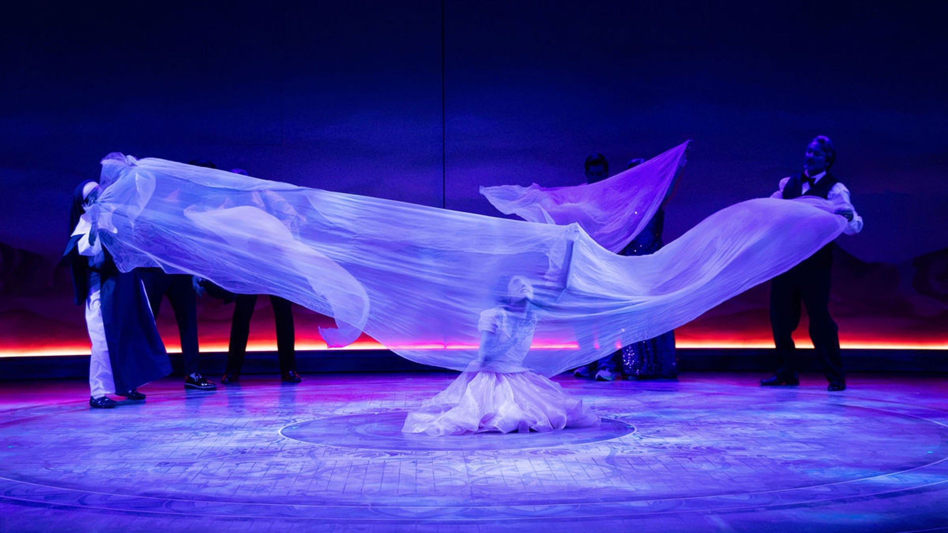 Actors stretch a long, sheer piece of fabric across the stage. An actor sits underneath it, reaching upwards.