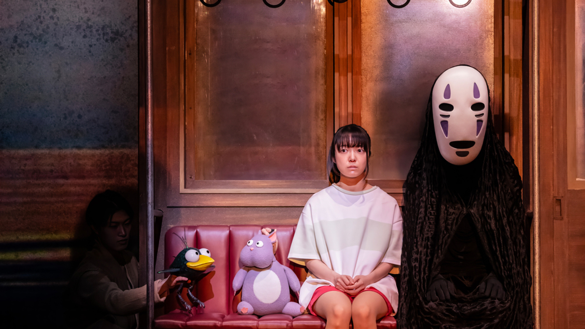Two actors sit side by side on a train facing the audience. The actor on the right is wearing a white mask and black robes. The backdrop behind them is a mixture of blue and yellow sky.