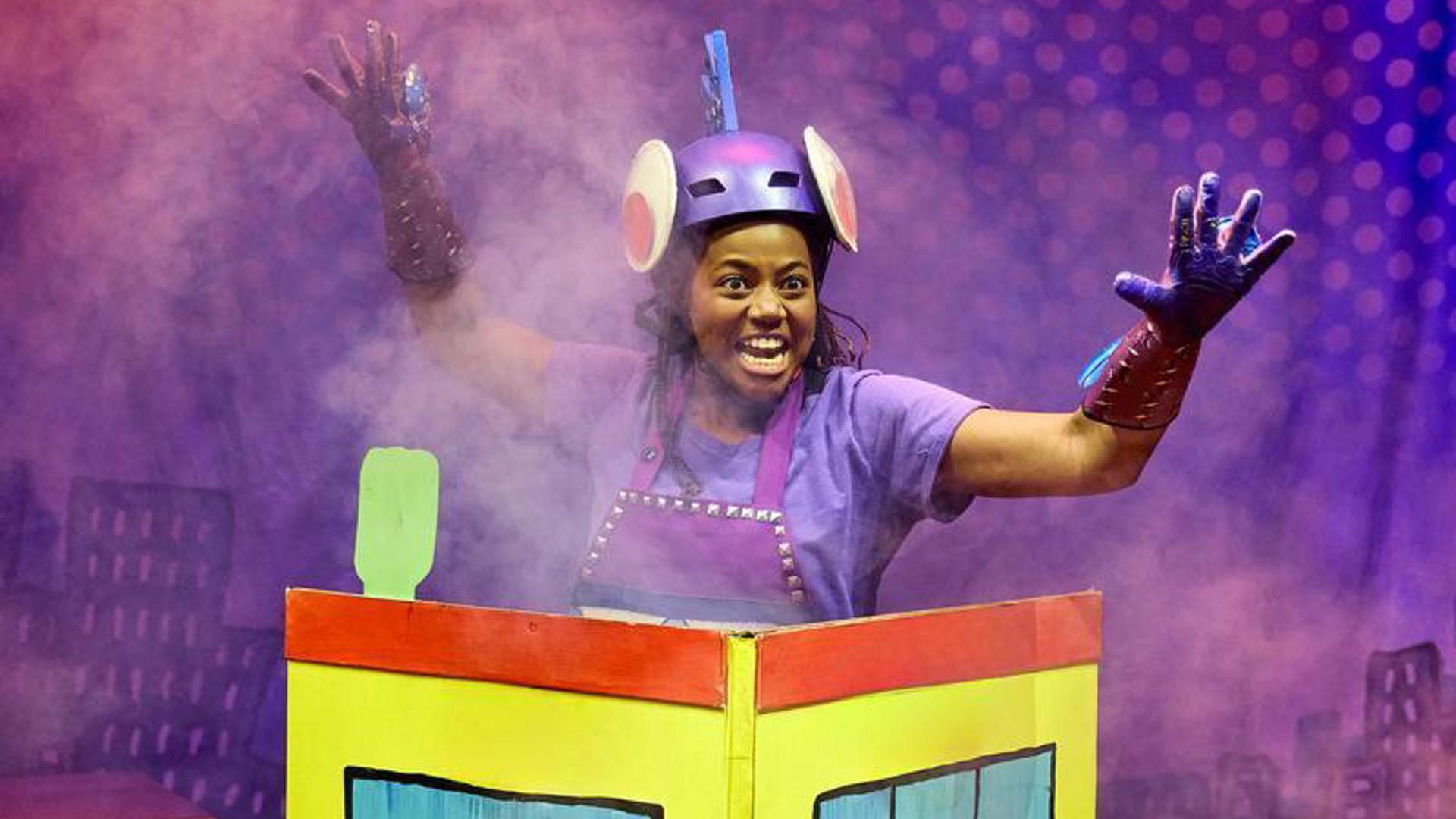 An actor emerges from the top of a cartoon building labeled "Living Spray Factory." There is smoke around the stage.