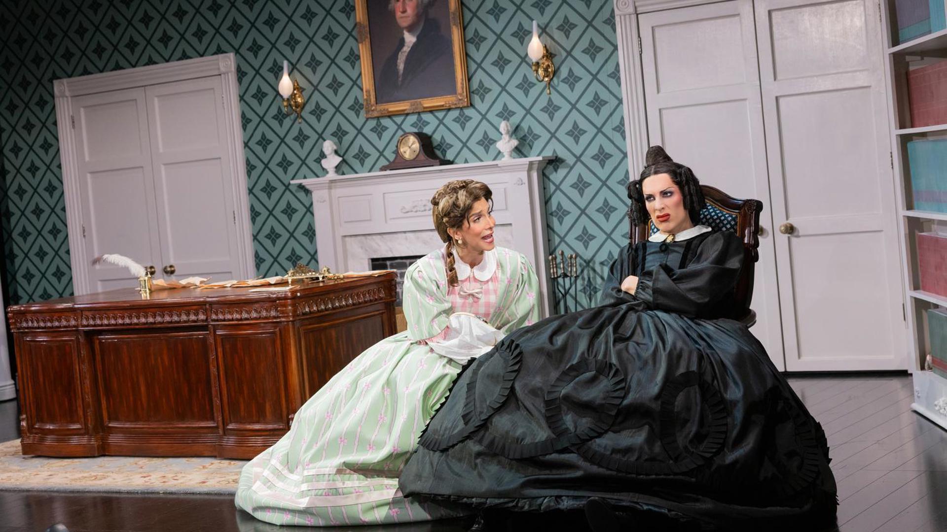 An actress in a black dress sits in a chair with a disgusted look on her face. To her right, an actress in a light green dress speaks to her.