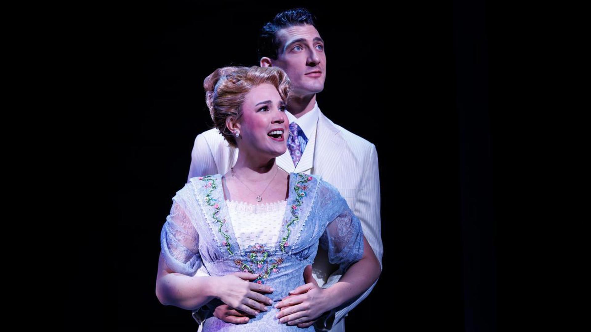 Two actors embrace with one standing behind the other, looking out at the audience. The actor in the back has his arms around the waist of the actor in front.
