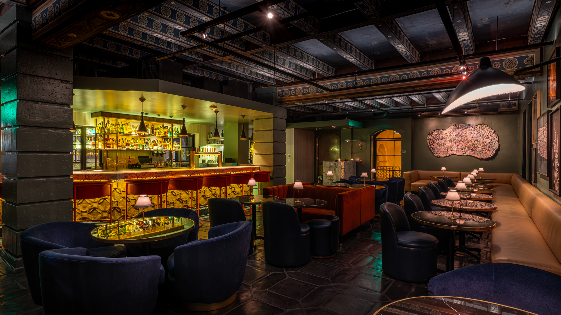 Moodily lit lounge bar with a marble-topped counter and back-bar shelves of liquor bottles, surrounded by navy velvet armchairs, an orange banquette, and a long tan booth seat along the wall, all set on dark octagonal floor tiles under a patterned ceiling.