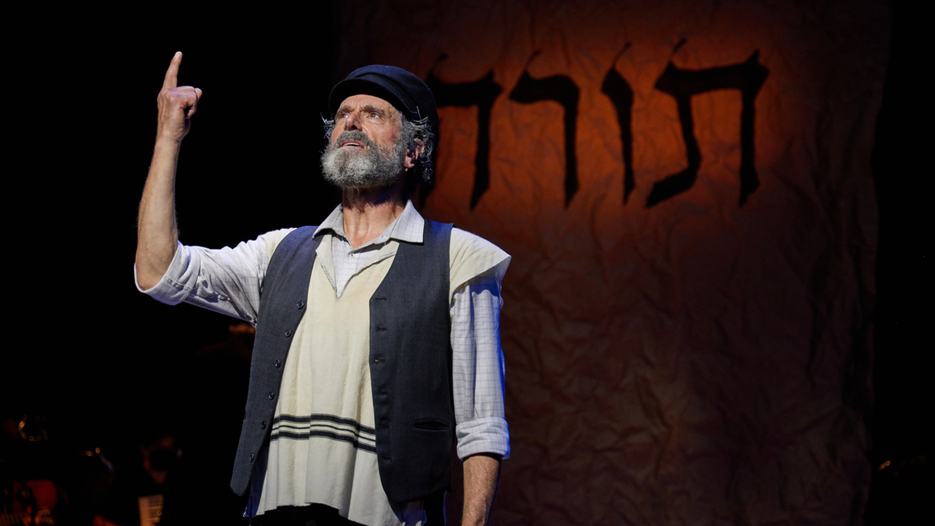 A man stands on stage stands and points his right arm up at the sky while looking up slightly. He is wearing a black hat and a gray vest as well as a tallit.