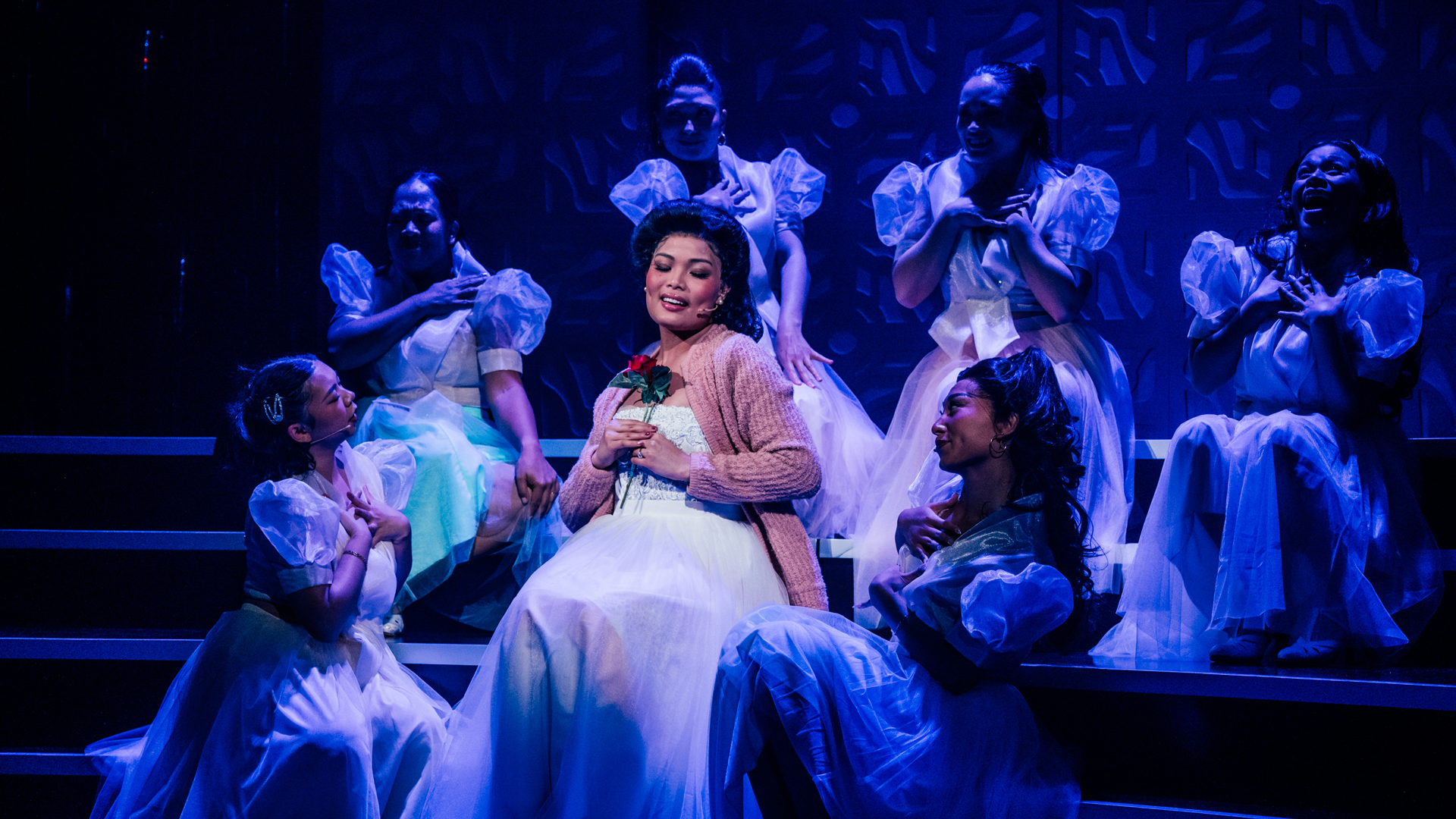 A performer sits, holding two roses to their chest, surrounded by dancers who clutch their hearts.