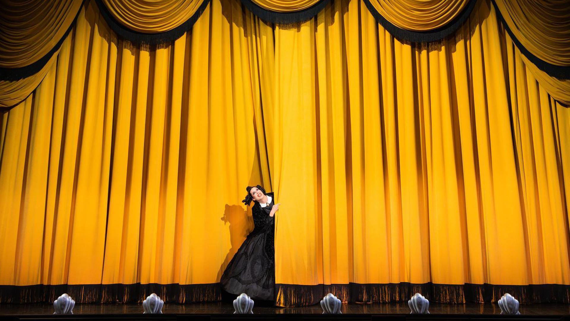 An actress in a black dress sticks out of a closed yellow stage curtain, smiling.