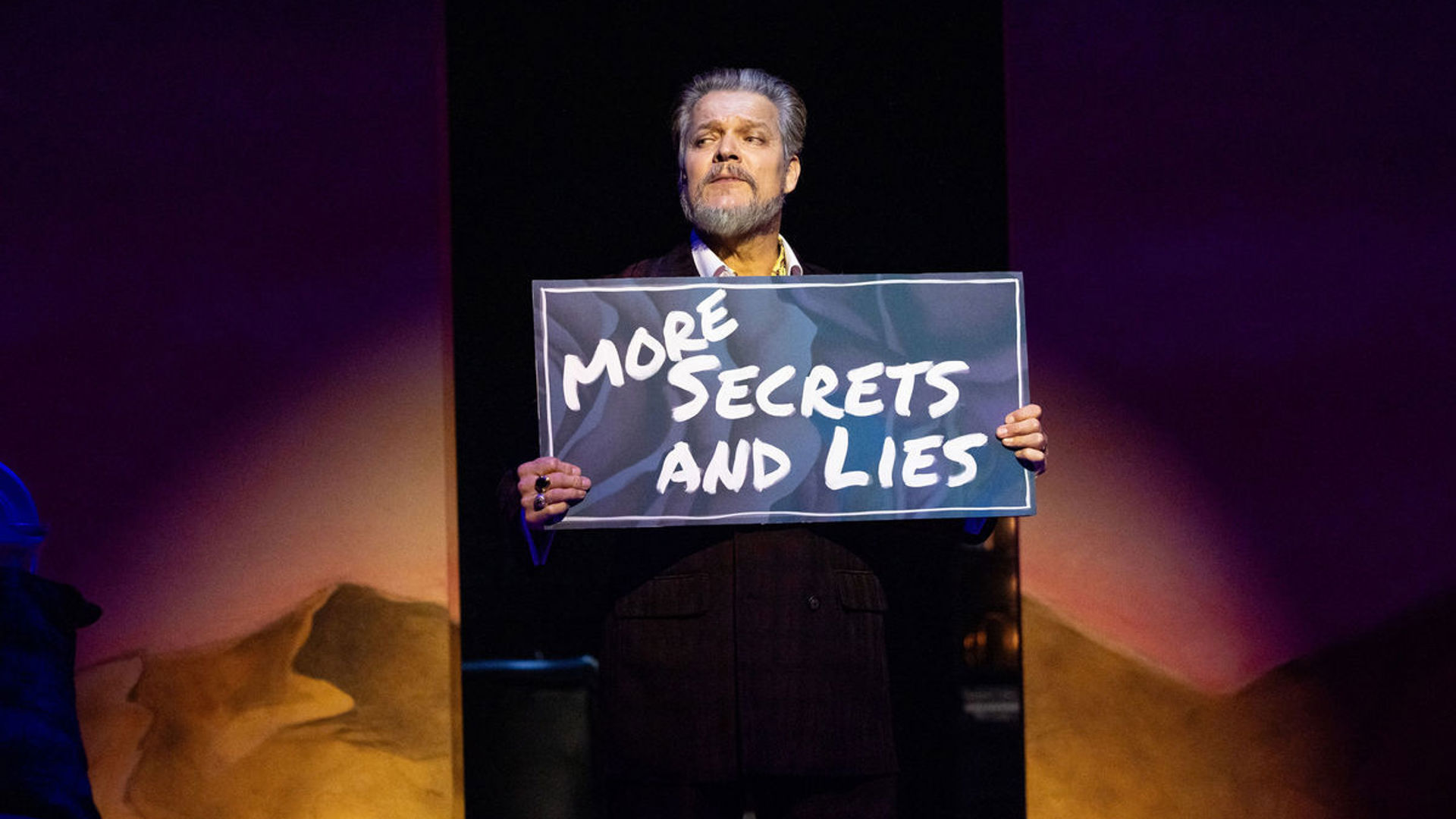 A man stands with a sign displaying "More Secrets and Lies"