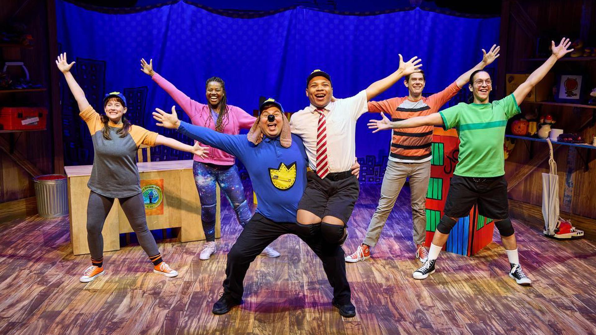 An actor dressed as dog man squats at center stage with their right arm extended, they are holding another actor on their hip, that actor has their left arm extended. Their are four actors behind them with both arms extended. They are all smiling.