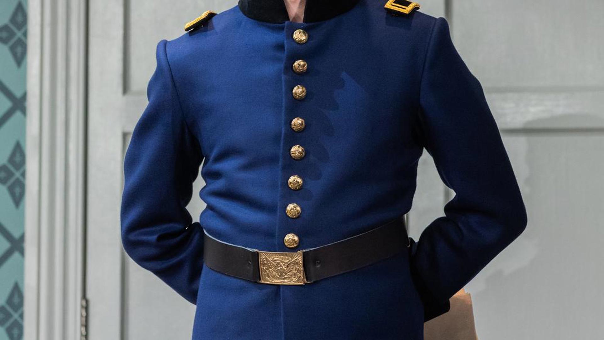 An actor stands in a blue soldiers outfit with his hands behind him, he looks out at the audience and smiles.