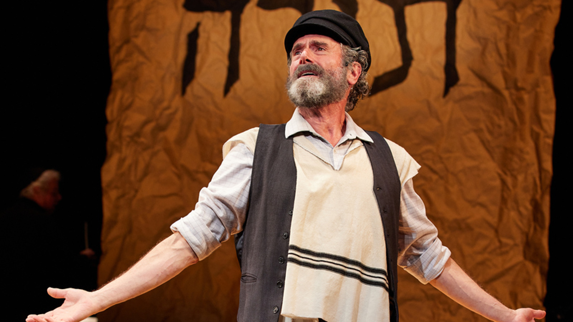 A man stands on stage stands with his arms out. He is wearing a black hat and a gray vest as well as a tallit. There is a jug to the right and behind him.