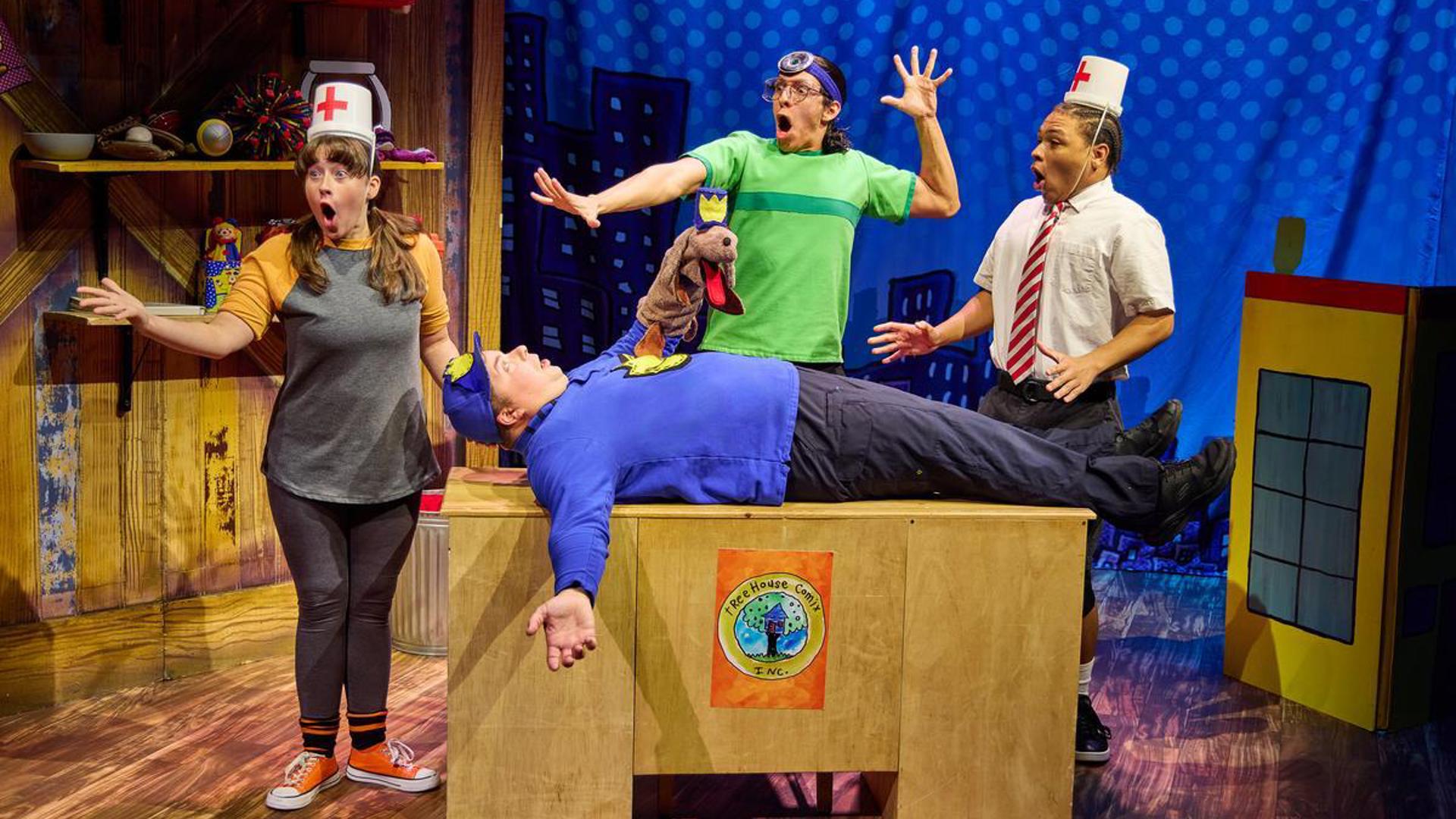 An actor lays on a wooden desk, staring up at the ceiling and holding a dog man puppet. Three actors stand around them dressed as doctors with their mouths agape.