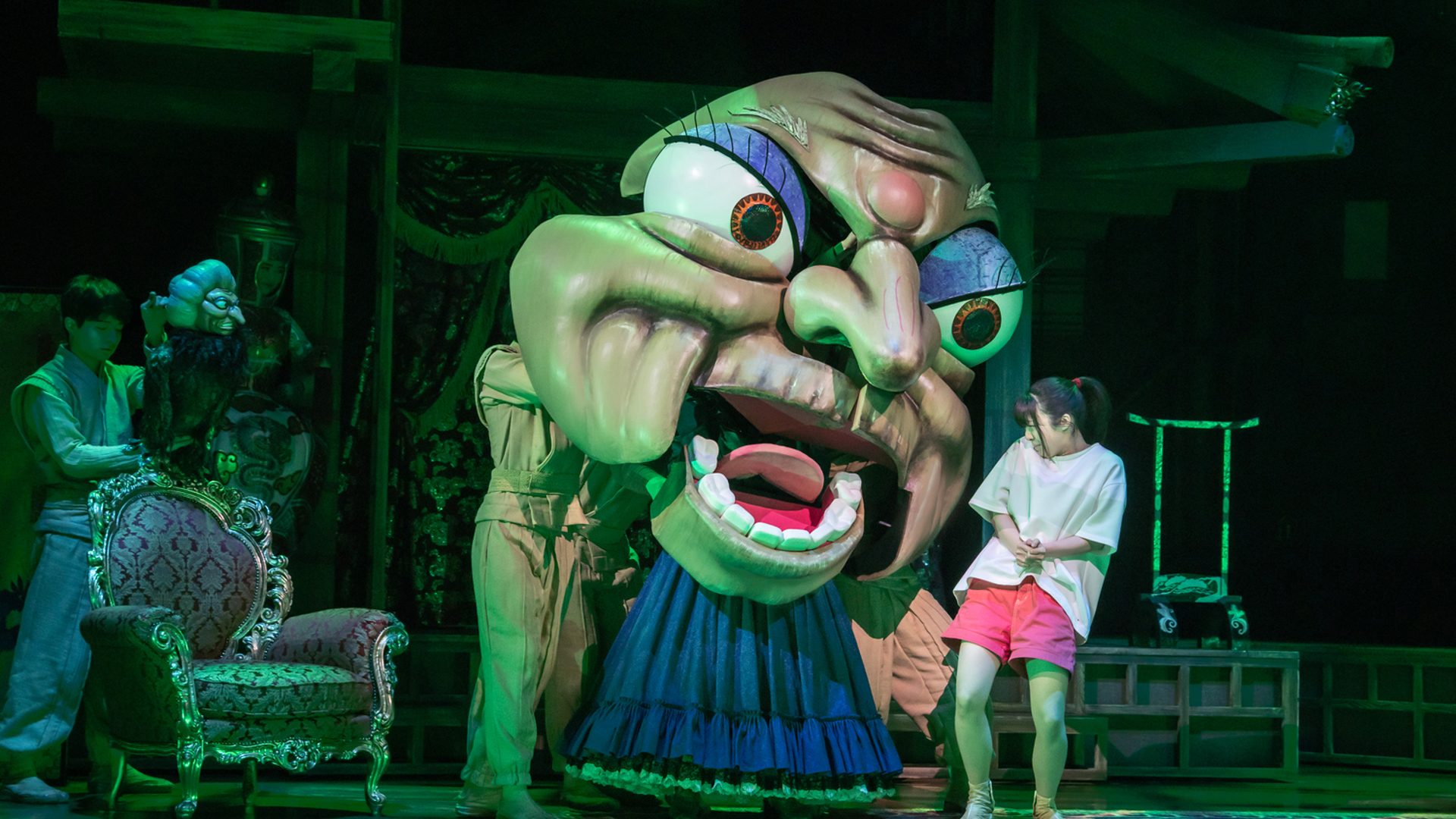 An actor cowers next to a large creatures face, looking at her intensely.