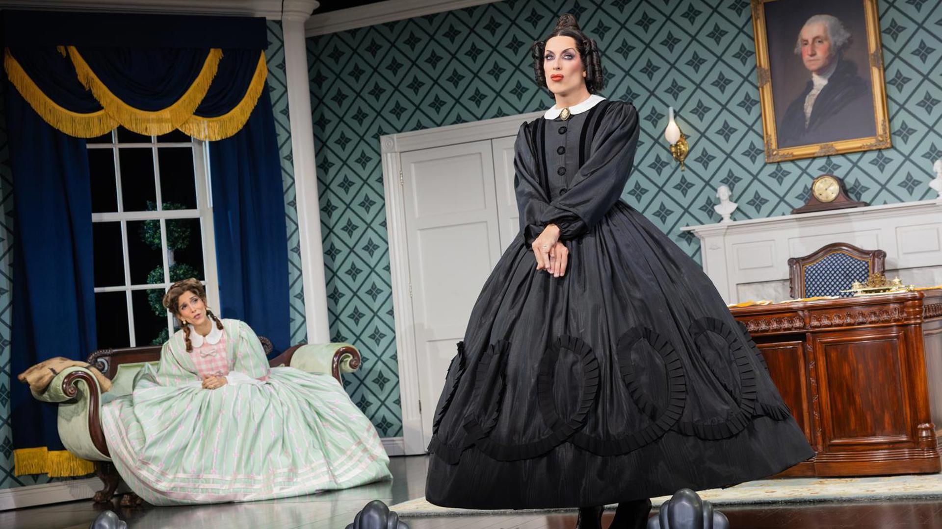 A woman in a black dress stands with her hands clasped in front of her, looking out at the audience. A woman in a green dress sits on a couch behind her, speaking to her.
