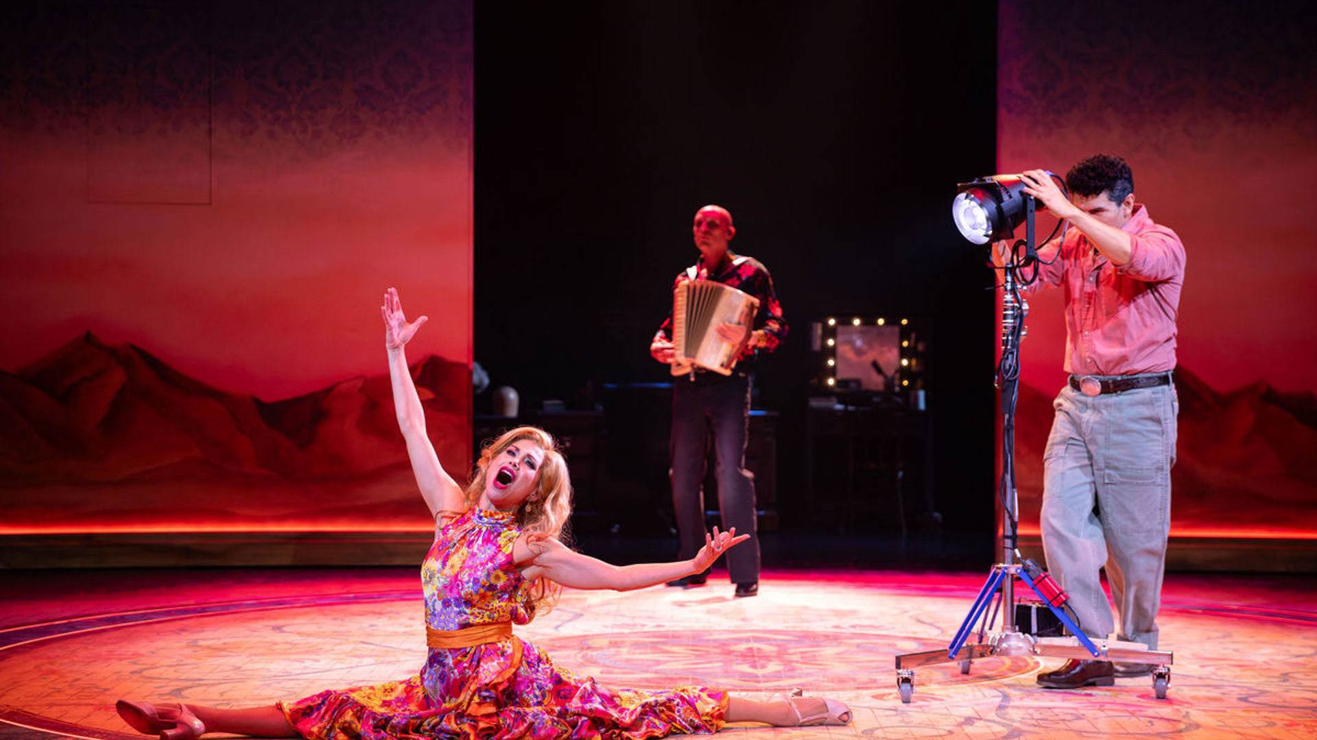 An actor does a split onstage, arms and mouth open as if singing. Behind her, an actor holds an accordion, and another actor holds a spotlight.