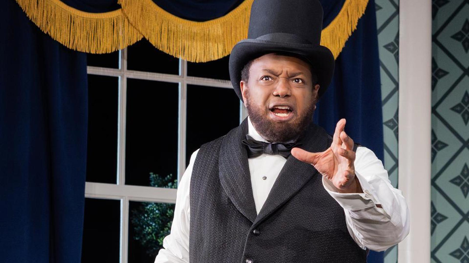 An actor stands in front of a window in a top hat. He is speaking to the audience and extending his left hand.