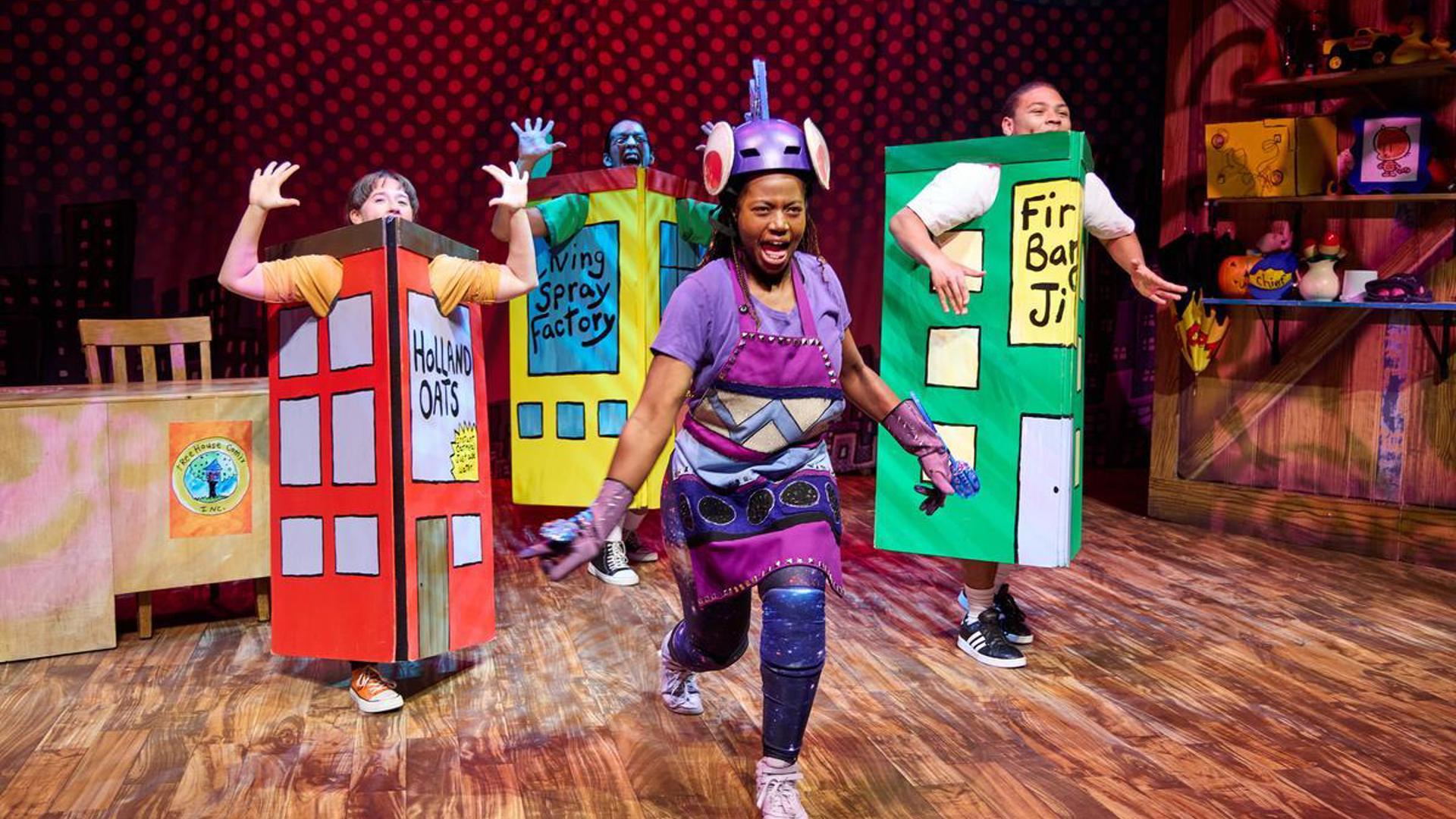 An actor lunges towards the audience with arms extended, dressed in a purple helmet, apron and galaxy leggings. Three actors stand around them in a semi-circle dressed as buildings.