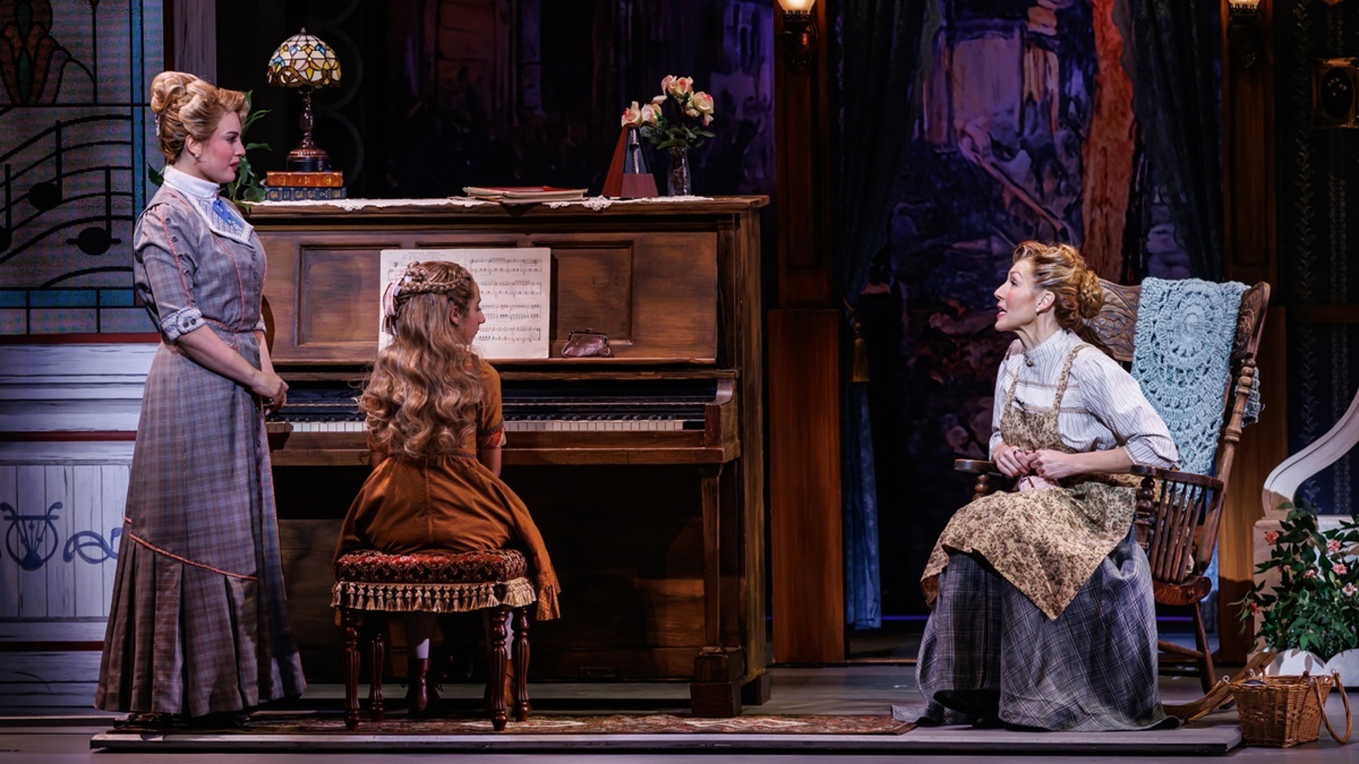 Two actors look at each other speaking, the one on the left is standing while the one on the right is sitting in a rocking chair. In between them, a young actor sits at a piano with her back to the audience.