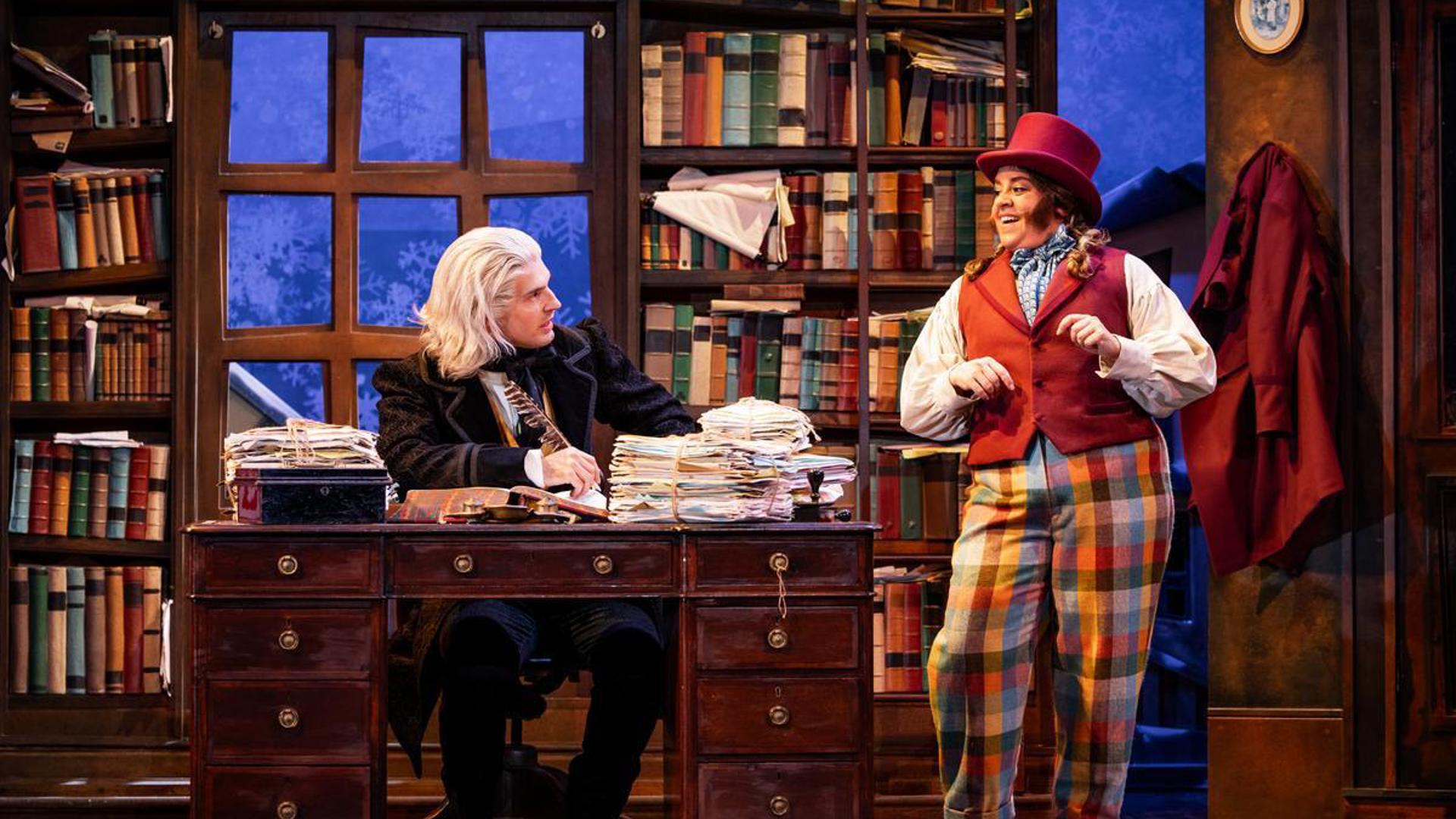 Two performers are side by side, one at a desk piled with papers and one standing next to the desk.