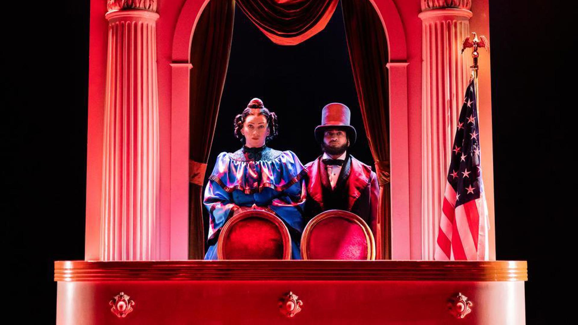 Two performers standing side by side directly behind chairs. There is an American flag to the right of them.