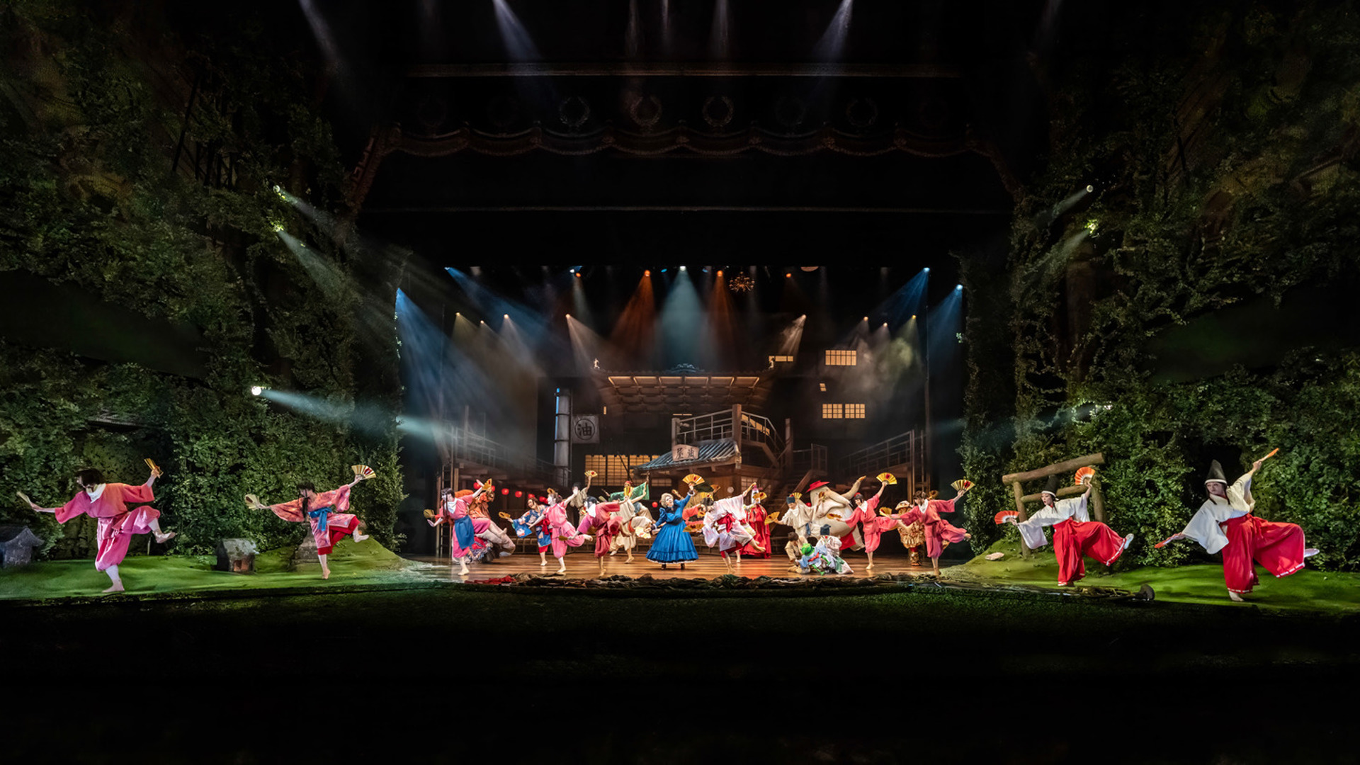 A large group of actors dance on stage, spread out across the stage. They have fans in each hand and are raising their left legs.