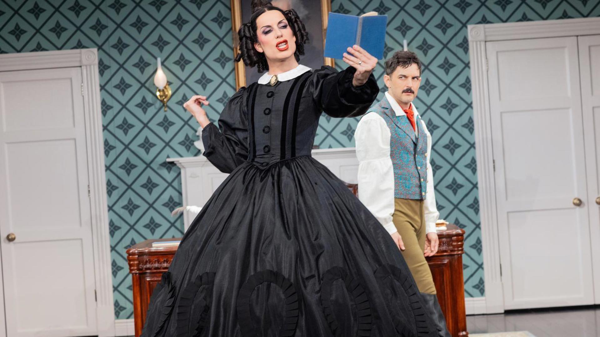 An actor stands in a black dress reading a book, another actor walks behind her looking at her.