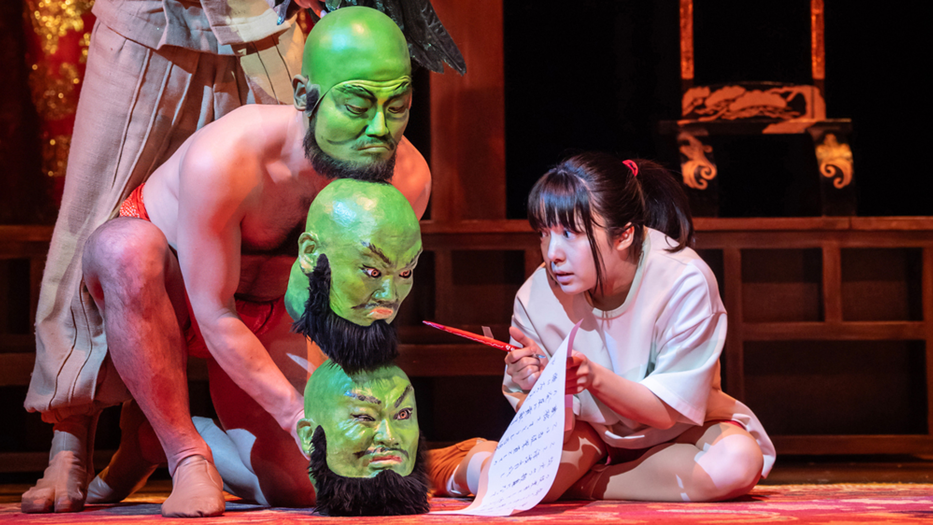 An actor crouches, holding a piece of paper and a brush and writing. Another actor crouches next to her, one controlling a puppet of two green heads stacked under his head, which is painted green.